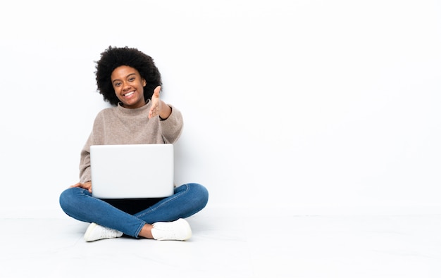 Young African American woman with a laptop sitting on the floor shaking hands for closing a good deal