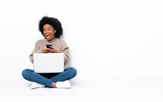 Young African American woman with a laptop sitting on the floor surprised and sending a message