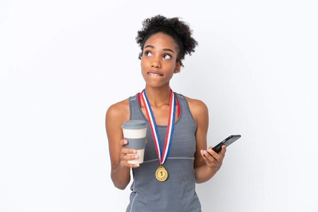 흰색 배경에 격리된 메달을 가진 젊은 아프리카계 미국인 여성은 커피를 들고 무언가를 생각하는 동안 휴대전화를 들고 있습니다