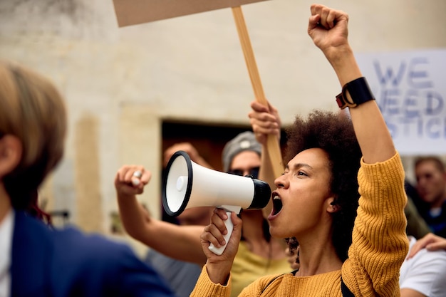 Young African American woman with raised fist shouting through megaphone while being on antiracism protest