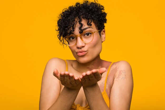 Young african american woman with skin birth mark folding lips and holding palms to send air kiss.