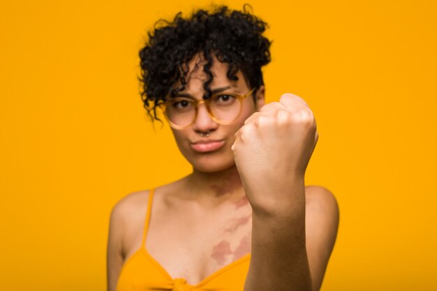 Giovane donna afroamericana con il segno di nascita della pelle che mostra pugno alla macchina fotografica, espressione facciale aggressiva.