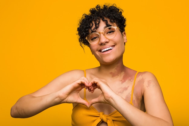 Young african american woman with skin birth mark smiling and showing a heart shape with hands