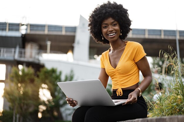 Молодая африканская американка, работающая на ноутбуке, сидит на скамейке в городе