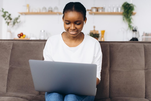 Young African American woman working on laptop sitting on sofa in kitchen