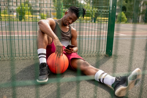 Photo young african basketball player with smartphone relaxing by fence of playground and scrolling in smartphone