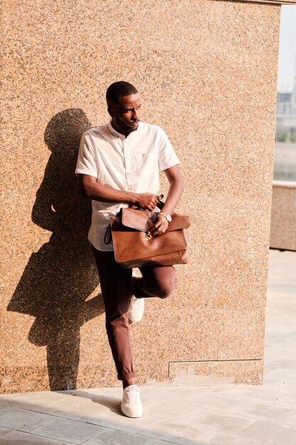 Young African businessman or agent in smart casualwear holding brown leather handbag on his knee while leaning against wall of building