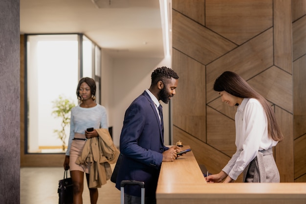 Foto giovane imprenditore africano in abiti da cerimonia chinandosi bancone della reception nella lounge dell'hotel mentre parla con l'addetto alla reception