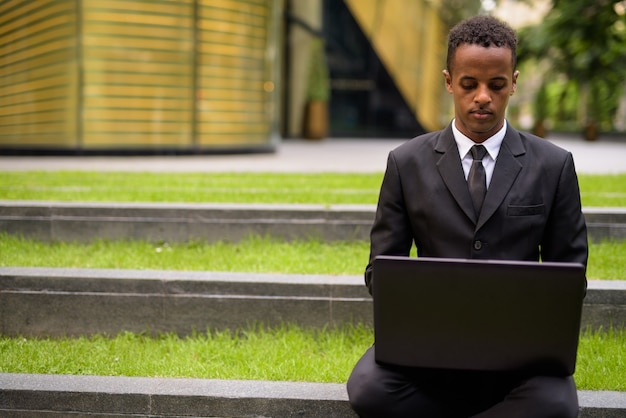 Young African businessman sitting and using laptop computer outdoors