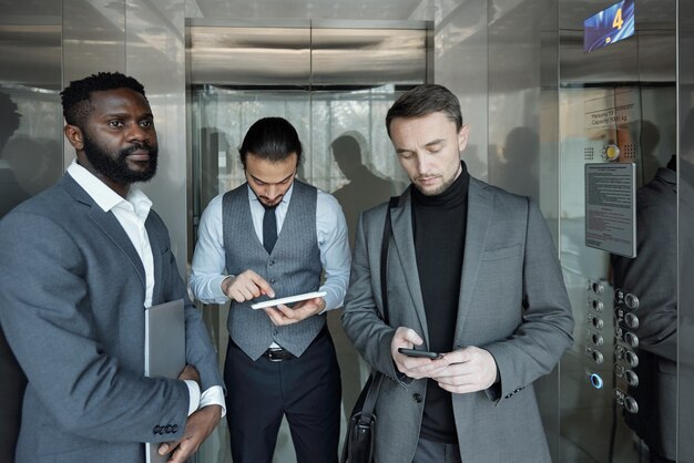 Young African businessman with folded laptop standing by wall of elevator while his two colleagues scrolling in tablet and mobile phone