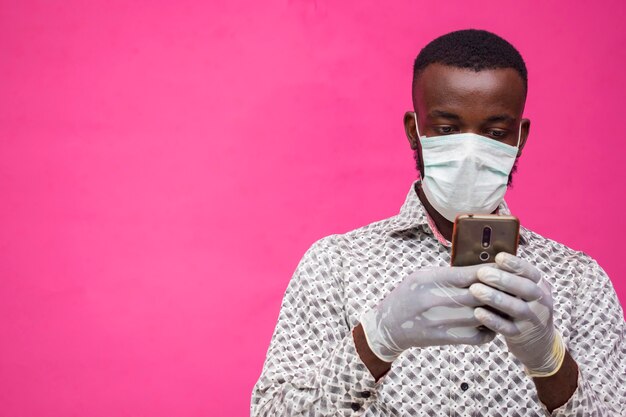 A young african doctor isolated over pink background wearing face mask to prevent, prevented, preventing himself from the outbreak in the society, operating his smartphone, telephone, mobile phone.