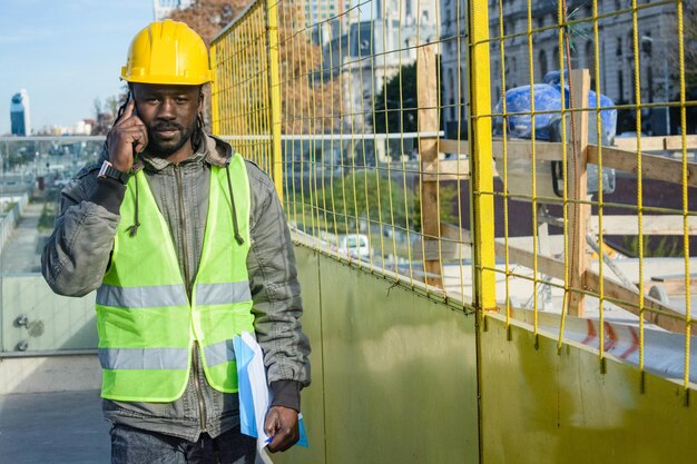 Молодой человек африканского происхождения, руководитель, разговаривает по телефону и гуляет по строительной площадке.