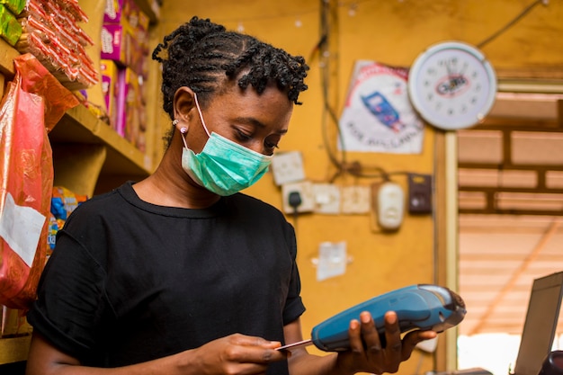 Young african female attendant using the point of sale machine to pay for the goods her customer bought while using face mask to prevent herself from the corona outbreak.