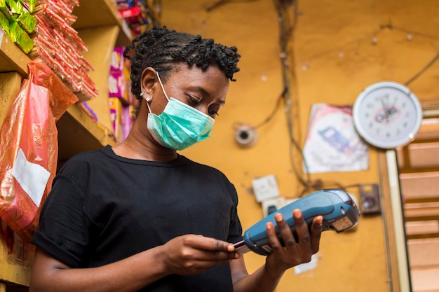 젊은 아프리카 여성 승무원은 코로나 발병을 예방하기 위해 안면 마스크를 사용하는 동안 고객이 구매한 상품에 대해 POS 기계를 사용하여 지불합니다.