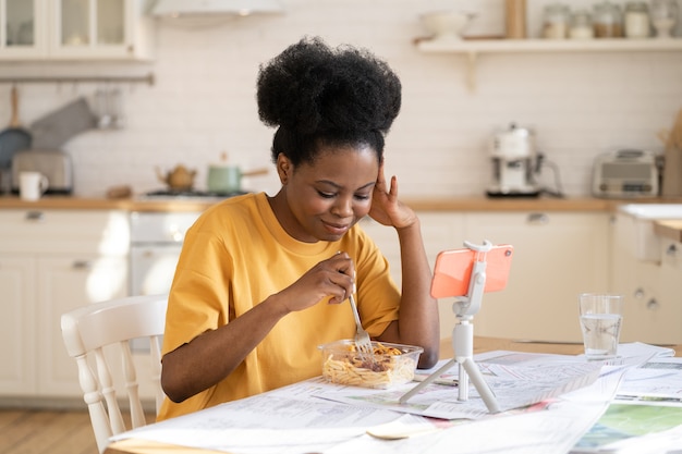 Young african female calling friends online via video chat in home kitchen at lunch break from study