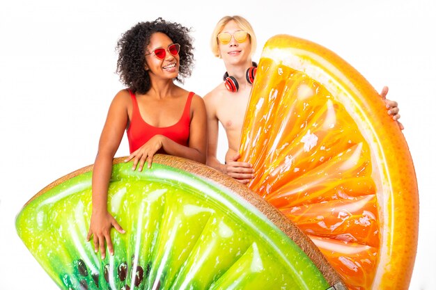 Young african female and caucasian blonde man stands in swimsuit with big rubber beach mattresses and smiles, isolated on white wall