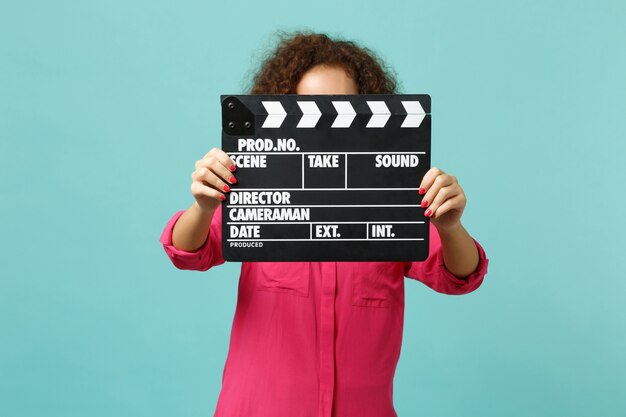 Young african girl in casual clothes covering face with classic black film making clapperboard isolated on blue turquoise background. People sincere emotions, lifestyle concept. Mock up copy space.