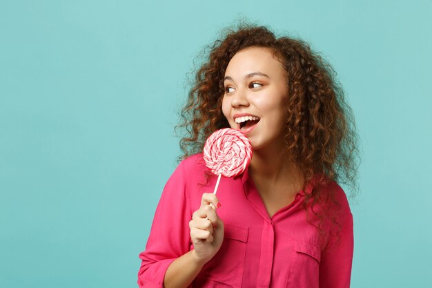 Photo young african girl in casual clothes holding, eating pink round lollipop, looking aside isolated on blue turquoise background in studio. people sincere emotions, lifestyle concept. mock up copy space.