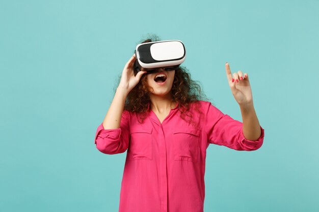 Young african girl looking in headset, touch something like push click on button, pointing at floating virtual screen isolated on blue turquoise background. People sincere emotions, lifestyle concept.