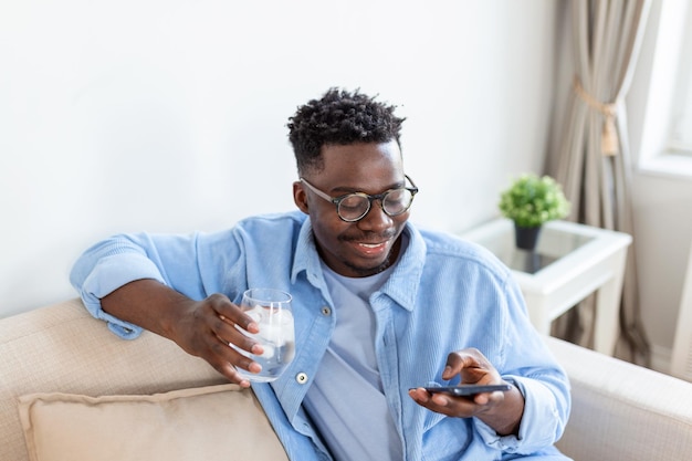 Young African man in casualwear drinking water while reading sms or dialing number on cellphone