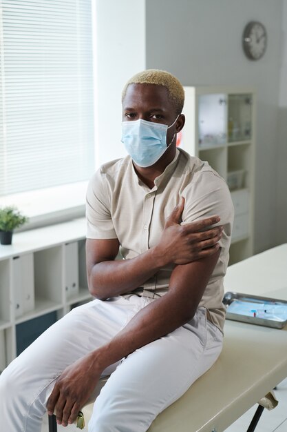 Young African man in casualwear and protective mask keeping hand on left shoulder after injection while sitting in front of camera in clinics