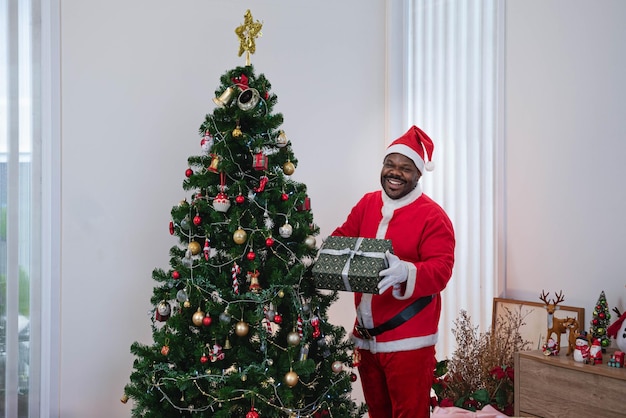 Giovane africano vestito in costume di babbo natale con una scatola regalo in piedi vicino all'albero di natale sorride felice a natale