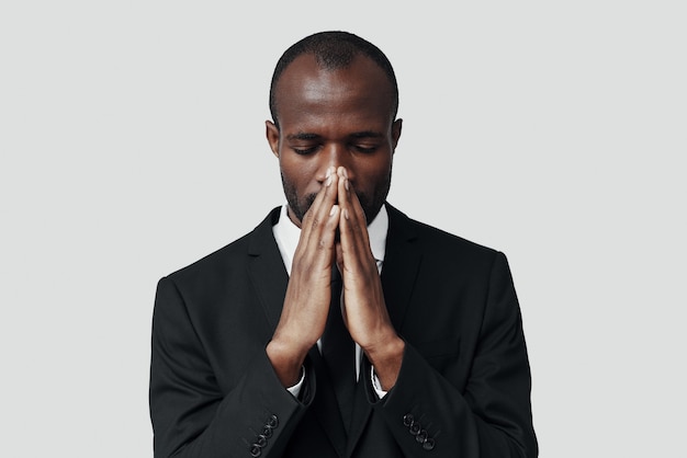 Young African man in formalwear keeping hands clasped while standing against grey wall