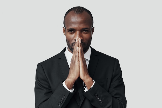 Young African man in formalwear keeping hands clasped while standing against grey wall