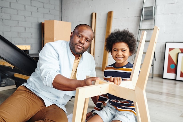 Young african man and his cheerful little son assembling wooden chair while sitting on the floor inside their new flat or house