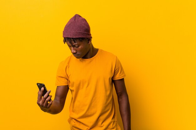 모자를 쓰고 전화를 사용하여 노란색 배경에 서있는 젊은 아프리카 남자