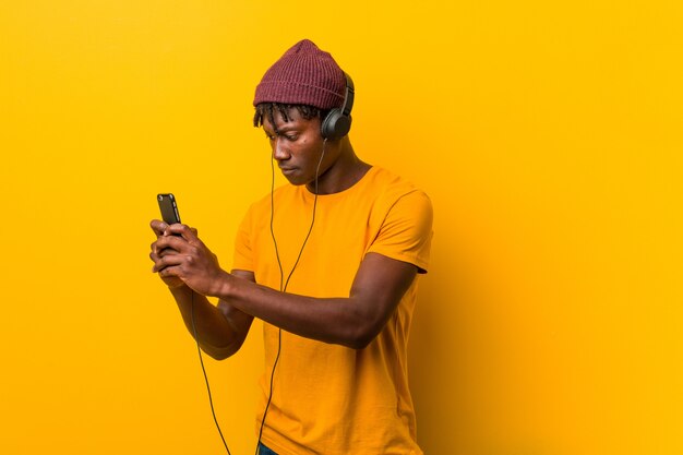 携帯電話で音楽を聞いて帽子をかぶって黄色に対して立っている若いアフリカ人