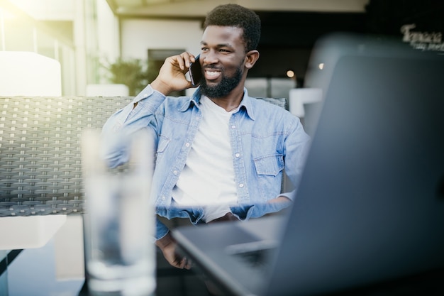 Young african man talking phone while sitting with laptop in cafe