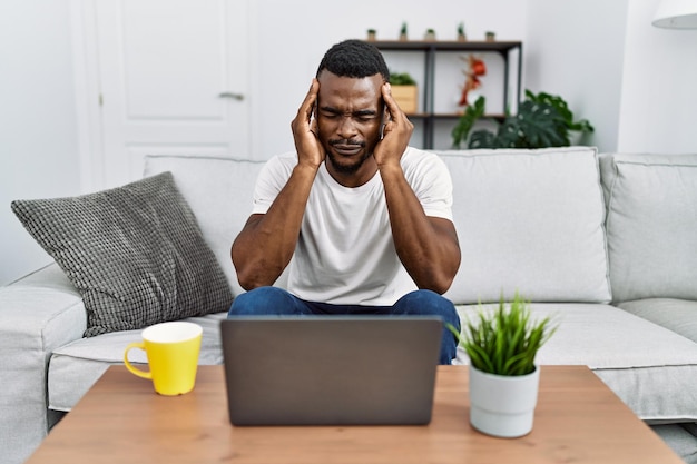 Young african man using laptop at home with hand on head for pain in head because stress. suffering migraine.