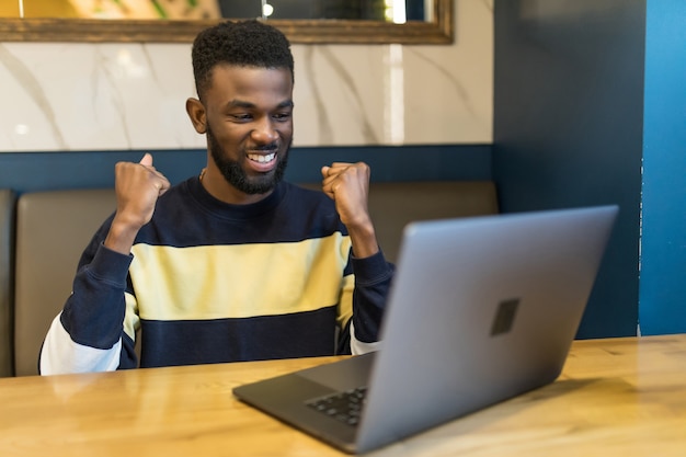 Young African Man with laptop Celebrating in a modern coffee shop