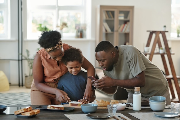 Photo young african man with smartphone taking photo of his wife and their cute little son cutting bread for sandwiches while preparing breakfast