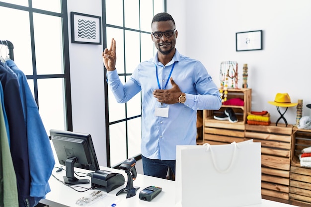 Young african man working as manager at retail boutique smiling swearing with hand on chest and fingers up making a loyalty promise oath