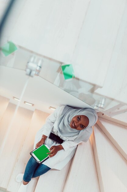 Young african modern muslim woman using tablet computer while sitting on the stairs at home wearing