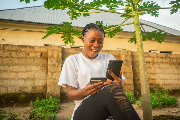 молодая африканская нигерийка с заплетенными волосами держит мобильный телефон и кредитную карту и делает покупки в Интернете