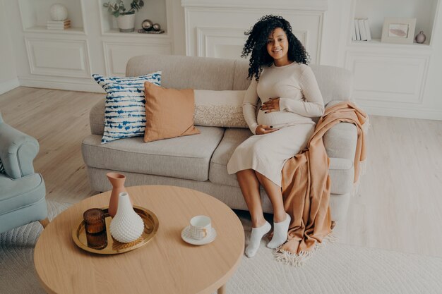 Young african pregnant woman in casual dress caressing her belly while sitting on couch at home