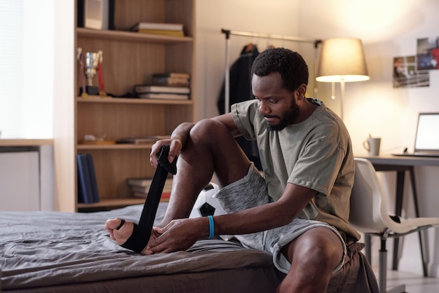 Young African sportsman putting black flexible bandage on his foot while sitting on bed and preparing for training