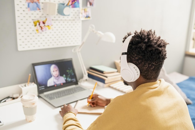 Young African student in headphones sitting in front of laptop with teacher on screen