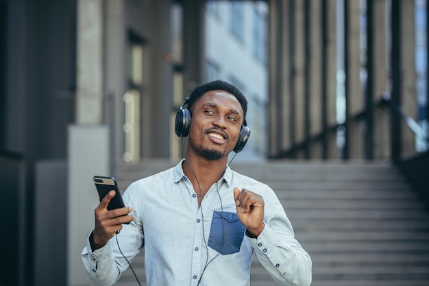 Young african student listening to music from smartphone using big headphones, smiling from the convenience of using the app