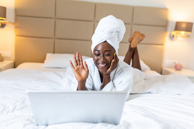 young African woman in bathrobe resting on bed after shower while working on her laptop in the morning.