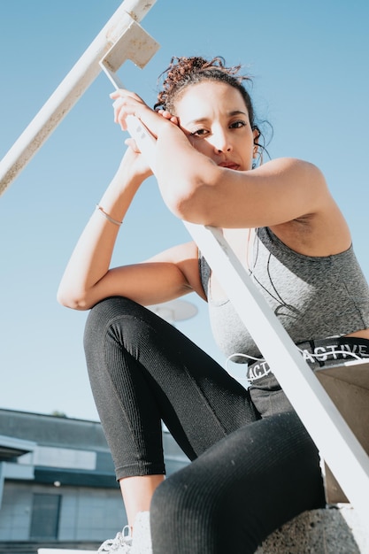 Foto giovane donna africana poc seduta su una sporgenza felice dopo aver fatto molto esercizio fisico e ascoltato musica, bella posa, con copia spazio, in leggins sportivi e top. ragazza in forma con i capelli ricci, bella donna