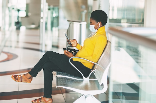 A young African woman in a protective mask sits in a chair in the waiting room, listening to music with headphones and waiting for a subway train. Social distance, pandemic.