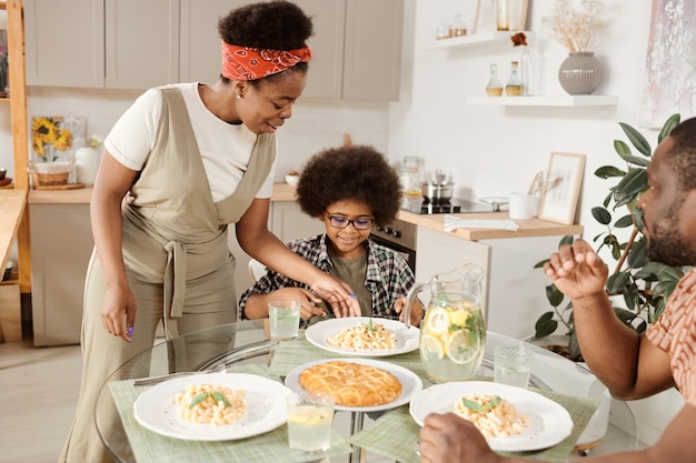 Young african woman putting plate with pasta on served table by her little son