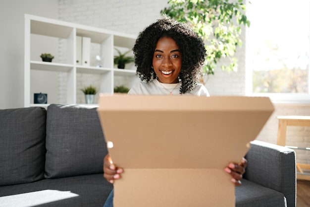 La giovane donna africana si siede sul divano a casa disimballando la scatola di cartone del pacco con l'acquisto online