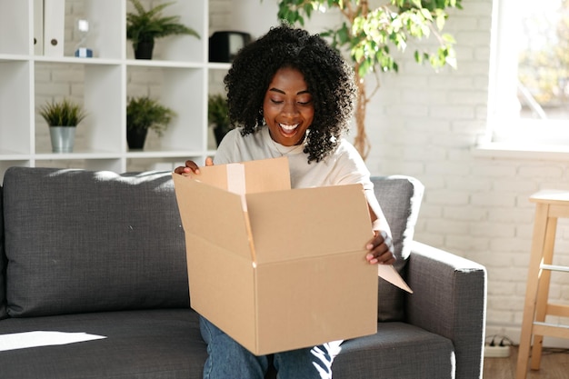 Young african woman sit on couch at home unpacking parcel cardboard box with online purchase