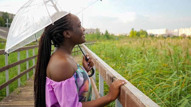 Young african woman under transparent umbrella in rainy weather on the bridge.