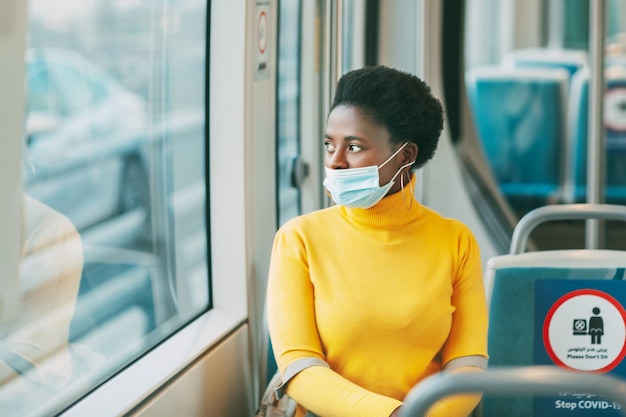 A young African woman wearing a protective mask rides on a bus and looks out the window. Coronavirus, social distance.